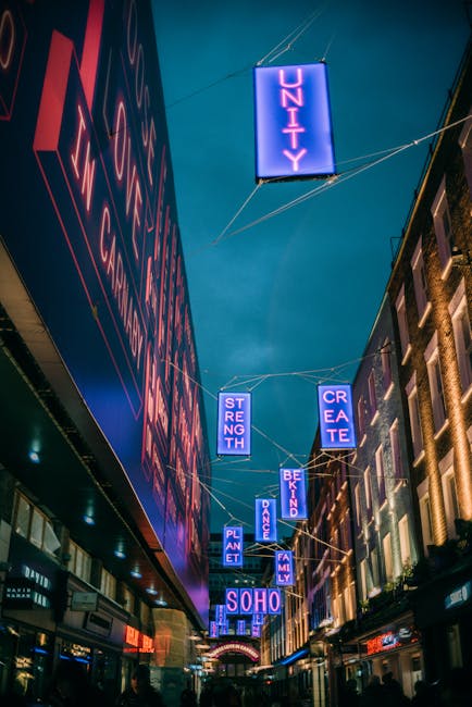 Nighttime street scene on Carnaby Street in Soho, illuminated by vibrant blue and purple neon signs displaying words such as 'UNITY', 'STRENGTH', 'CREATE', 'PLANET', 'DANCE', 'FAMILY', and 'BE KIND'. The signs are suspended above the busy pedestrian walkway by thin wires, with storefronts lining both sides of the street, some with illuminated signs and window displays. The sky above is dark, contrasting with the bright neon lights, and the scene reflects the lively atmosphere typical of central London areas. In the context of Soho removals, this image illustrates the urban environment where furniture and belongings may be transported during home relocation or moving services, with a focus on the vibrant district's commercial and cultural atmosphere.
