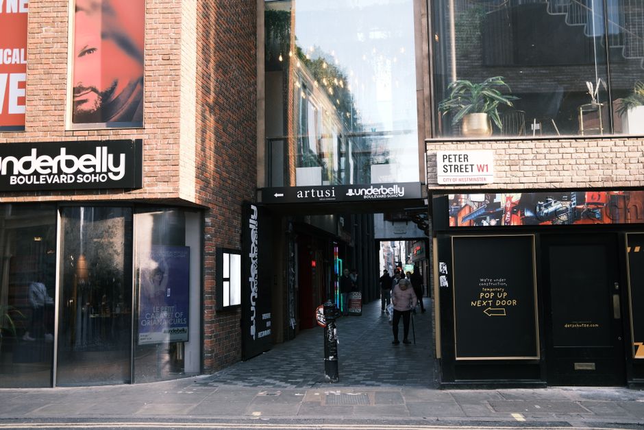 An urban street scene showing the entrance to a Soho building on Carnaby Street with a black sign displaying 'artusi' above a narrow passageway used for home relocation activities. To the left, there is a brick facade with a large window and a sign reading 'underbelly' for a bar or venue, while on the right side, there is a black storefront with yellow text indicating 'pop up next door'. In front of the entrance, pedestrians are walking along a paved sidewalk, some carrying bags. A black bollard and a bicycle are positioned near the doorway, which is part of the moving process involving furniture transport and packing and moving logistics. The environment is illuminated with natural daylight, reflecting urban activity typical of Soho, London, relevant for professional removals or house removals services provided by Soho Movers. The scene captures the typical exterior of a commercial property involved in a local moving or relocation activity, with clues of ongoing access or loading procedures inside the passageway.