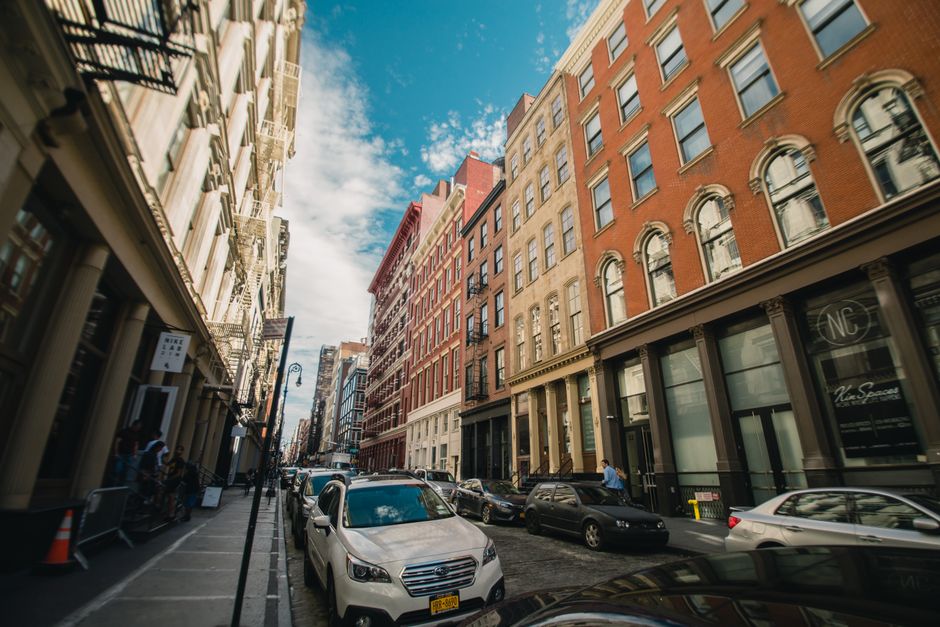 A view of a narrow street in Soho, W1, during daytime with modern and historic buildings on either side, featuring brick facades and large windows. The street is paved with dark bricks and lined with 