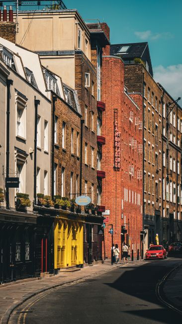 The image depicts a street scene in Soho with multi-storey brick buildings lining the narrow pavement, under a clear blue sky. In the foreground, a black and yellow moving trolley is seen loaded with 