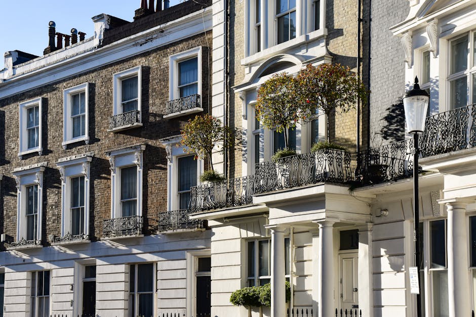 A row of narrow, multi-story residential buildings on a cobblestone street, featuring various exterior wall colours including white, black, and light blue. Each property has potted plants and small ga